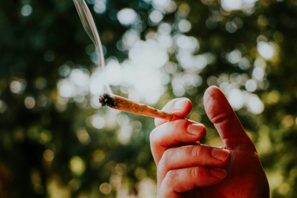 Hand holding a lit joint with smoke, outdoors, blurred background.